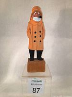 Front angled view of hand-carved wooden fisherman figurine in orange raincoat, black boots, with pipe and white beard.