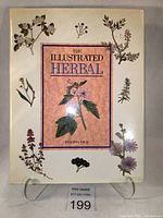 Front cover of The Illustrated Herbal by Philippa Back, showing illustrated herbs on dust jacket and book standing on display stand with lot tag visible.