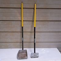 Full view of two metal dog poop scoops with yellow grips and long handles against a beige background.