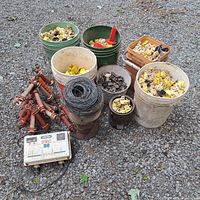 Wide photo showing all items laid out including electric fence controller, multiple buckets of yellow and black insulators, two red fence handles, coil of wire, clips and fittings.