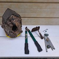 Four items on a white table: homemade bark bird house with peaked wood roof, green handled Schmidt loppers, small garden hoe with wooden handle and rusted blade, metal wind chime with tubes and green decorative balls.