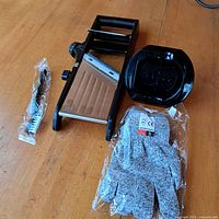 View of mandolin slicer, plastic food holder, cleaning brush, and packaged cut-resistant glove on wooden surface.