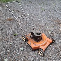 Side view showing the orange metal lawn mower deck, black engine, and metal handle over a gravel surface