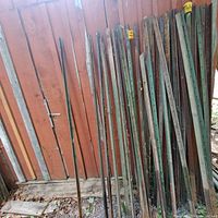 Row of 31 metal T-posts leaning against a shed wall; various paint and rust conditions visible, some topped with yellow caps.