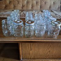 View of 18 assorted glass preserving jars, some with metal lids, arranged in rows on a wooden table.