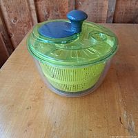 Salad spinner with green basket, clear container and green lid with blue knob for spinning. Viewed from the side on wooden table.