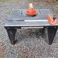 Front angle view of the Sears Craftsman router table showing aluminum grooved work surface, orange plastic fence attachment in middle, and orange guard piece on the right side. Black metal legs with rust and wear visible.