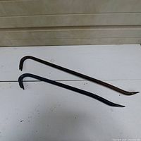 Two crowbars displayed on a white table against a beige wall, showing length and condition.