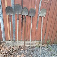 Five shovels standing upright against a wooden fence, different blade shapes and long wooden handles visible.