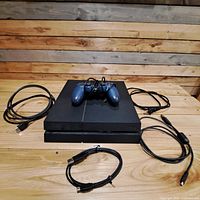 Sony PS4 console with blue controller on top and surrounding cables including HDMI, power, and USB charging cable.