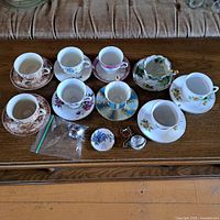 Photo of 7 teacups with matching saucers in different floral and scenic patterns alongside 2 metal tea infusers and 1 floral button pin