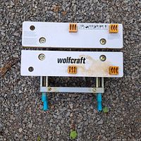 Top view showing two parallel white metal clamp plates with 'wolfcraft' printed on one, orange plastic grips, and visible screw mechanisms with blue plastic handles.