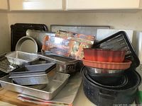 Various bakeware and roasting pans stacked including metal loaf pans, cookie sheets, pie plates, cooking racks, along with grill mats and a wood cutting board.