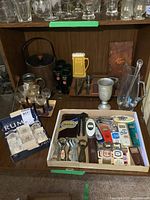 Wide view showing assorted glassware, stein, metal ice bucket, bottle openers and other bar tools in a wooden cabinet.