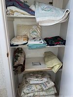 Shelving closet with folded bedding including quilts, sheets, runners, table cloths, and place mats in various colors and patterns