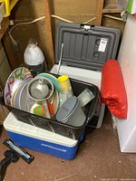Photo of blue Coleman cooler with black basket containing plastic plates, metal canteen, plastic cups and containers, next to gray powered cooler with lid open.