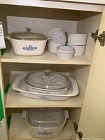 Cabinet shelf with three sizes of covered Corningware casseroles and six white ceramic ramekins.