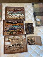 Multiple boxes containing a large assortment of costume jewelry including beaded necklaces, bracelets, rings, brooches, bolo ties, and a metal chain belt arranged on a mattress.
