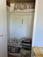 Photo of closet shelf and floor area showing large collection of crafting magazines in labeled white cardboard holders on upper shelf and lower shelf. Miscellaneous crafting supplies including beads, buttons, thread, and small plastic drawers are on the floor below.