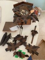 Full view of disassembled cuckoo clock showing carved wooden leaves, two birds, clock face with Roman numerals, and wooden tray.