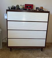Front view of the dresser showcasing four white slatted drawers and wooden frame with angled legs.