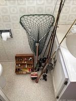 Fishing poles, fishing net, wooden tackle box with lures arranged on floor near bathroom sink and toilet