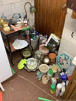 Wide view showing metal side table holding several ceramic planters, frog garden decor, glass balls, and stepping stones near a basement door.