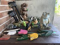 Photo showing large frog statues, solar stake lights, small frog figurines, wooden tulip stakes in multiple colors, and seashells arranged on a table outdoors.