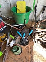 Photo showing green lawn seed spreader with metal handles, large plastic planter, yellow knee pad, hand gardening tools with blue and green handles, green ceramic planter, and rustic metal garden rake.