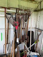 Photo showing a variety of garden tools including rakes, shovels, and a pitchfork all leaning against a shed wall.