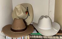 Three cowboy hats on a shelf: one straw with feather, one tan wool with braided band, one white wool with white band.