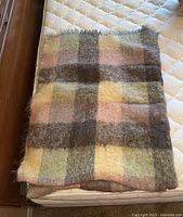 Folded mohair blanket on mattress showing plaid pattern with shades of brown, yellow, green, and pink.