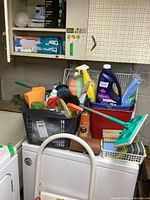 Photo showing cleaning supplies arranged in baskets, including Swiffer mop, spray bottles, detergent, brushes, sponges, buckets, and light bulbs above washing machines.