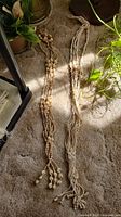 Photo showing two macrame plant hangers with knotted designs and wooden beads placed on a carpet, partial view of plants and pots around.