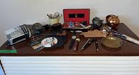 Wide view of men's accessories including watches, pipe, metal piggy banks, pocket change dish, spoon clackers, and alarm clock grouped on a wooden surface.