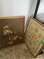 Photo of two framed needle art pieces: a floral embroidery with monarch butterfly and a cross stitched sampler, leaning against a wall.