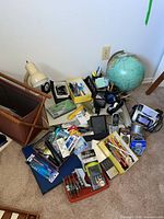 Photo showing the lot with a blue and green desk globe, beige desk lamp, brown leather magazine rack, various pens, writing utensils, rulers, batteries, a vintage Studebaker transistor radio, and other electronic and office items on carpet.