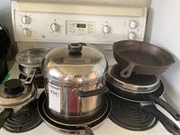 Cookware displayed on stove top including sauce pans with lids, mixed fry pans, cast iron fry pan, and stock pot with lid.