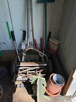 Overview of push reel lawn mower, garden tools including clippers and weed puller, terra cotta pots, garden fencing, garden hose nozzle, tomato fertilizer container in shed corner.