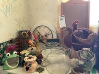 Photo showing assortment of glass bowls including rose-shaped, frog figurines, ceramic honey jar, bee figurine, wicker duck and chicken baskets, and battery clock in background