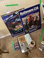 Overview photo showing two Rothmans racing posters leaning against the wall sized about 19x26 inches, several Benson & Hedges cigarette packs, matchboxes, tape, coasters, a branded mug, and other assorted collectibles arranged on the floor.