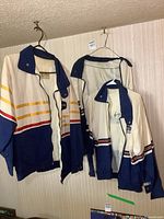 Three vintage Rothmans racing jackets hanging on wall, showing full view with distinct white, navy blue, and stripes pattern.