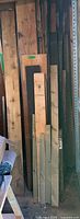 Photo showing multiple wood planks of different widths and lengths leaning against a shelf or wall, some stained or marked, length around 2-6 feet.