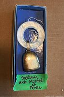 Baby rattle in box showing silver bell and mother of pearl ring with green sticker label.
