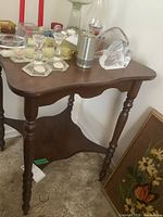 Wooden side table with turned legs and lower shelf shown at an angle with some items on top including glassware (not included). Table dimensions approximately 22x12x28 inches. Needs cleaning.