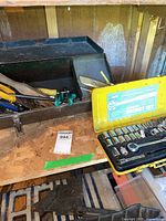 Metal toolbox with assorted hand tools including snips and carpet cutter beside a 40-piece ratchet socket set in a yellow case