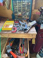 Woodworking bench with wood engraving chisels in yellow case, assorted C clamps, a hardware organizer with 26 cubbies filled with screws and hinges, snips, oil can, and other hand tools spread on bench and in drawer below.