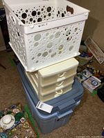Stack of storage containers including a white plastic basket with circular and paw print cutouts on top, a small craft organizer with 3 drawers below it, and two large storage bins beneath.