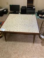 Front view of vintage coffee table showing full tiled square top and wooden legs on carpeted floor.