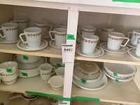 Photo showing multiple shelves holding white opaque glass dishes with gold-brown floral trim, including mugs, plates, bowls, cream and sugar set.